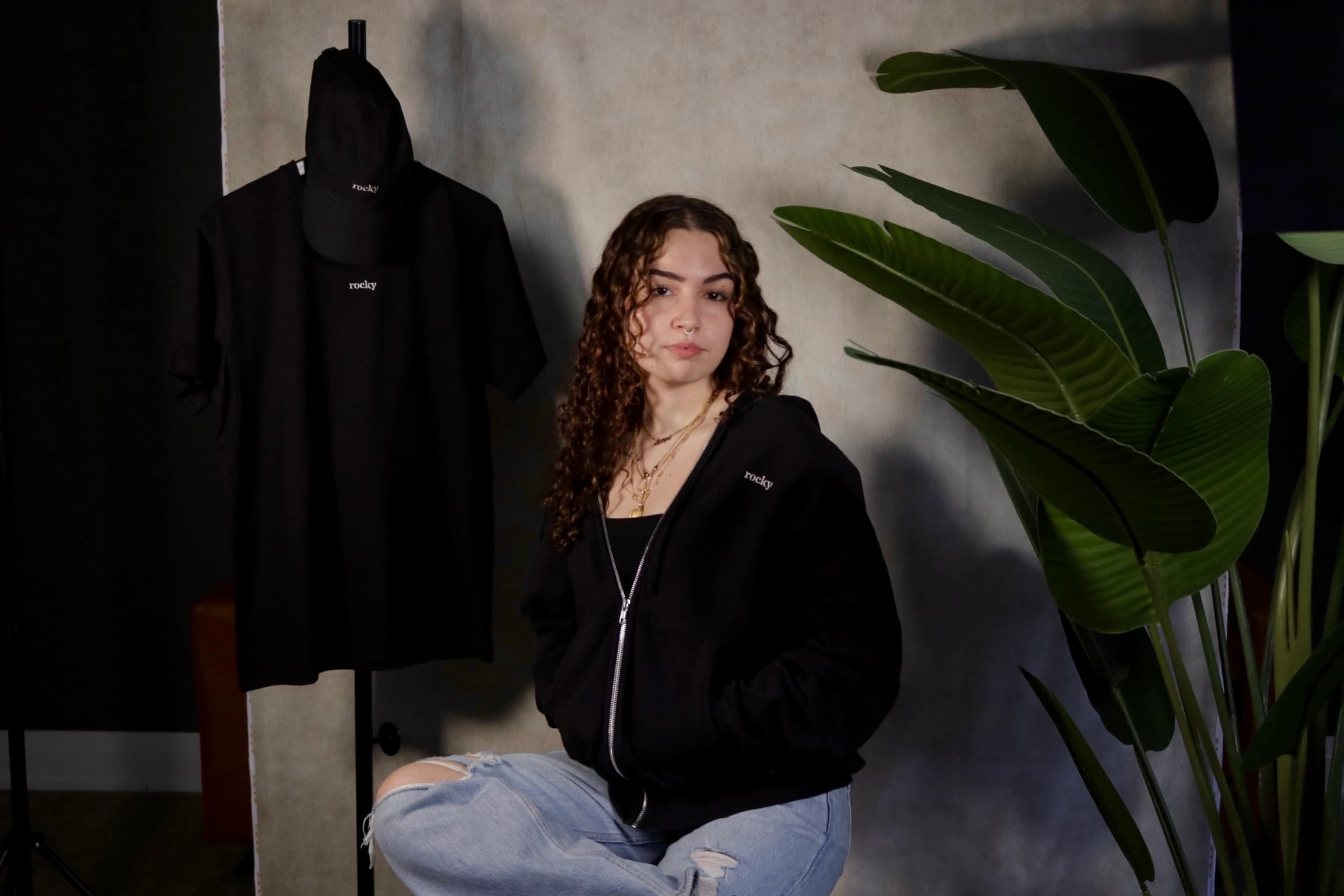 Person with curly hair wearing black top with green plants background