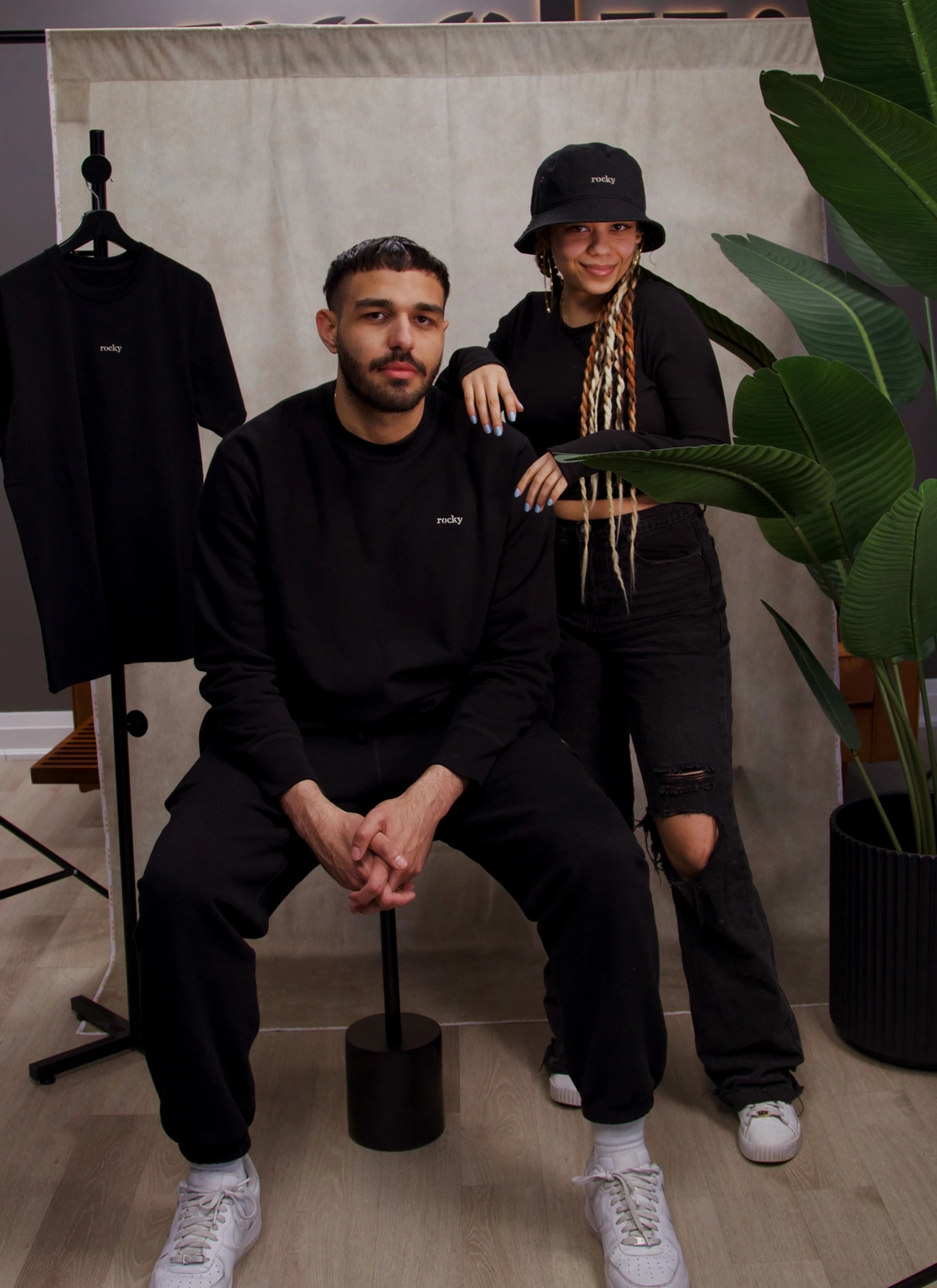 Person with curly hair wearing black top with green plants background