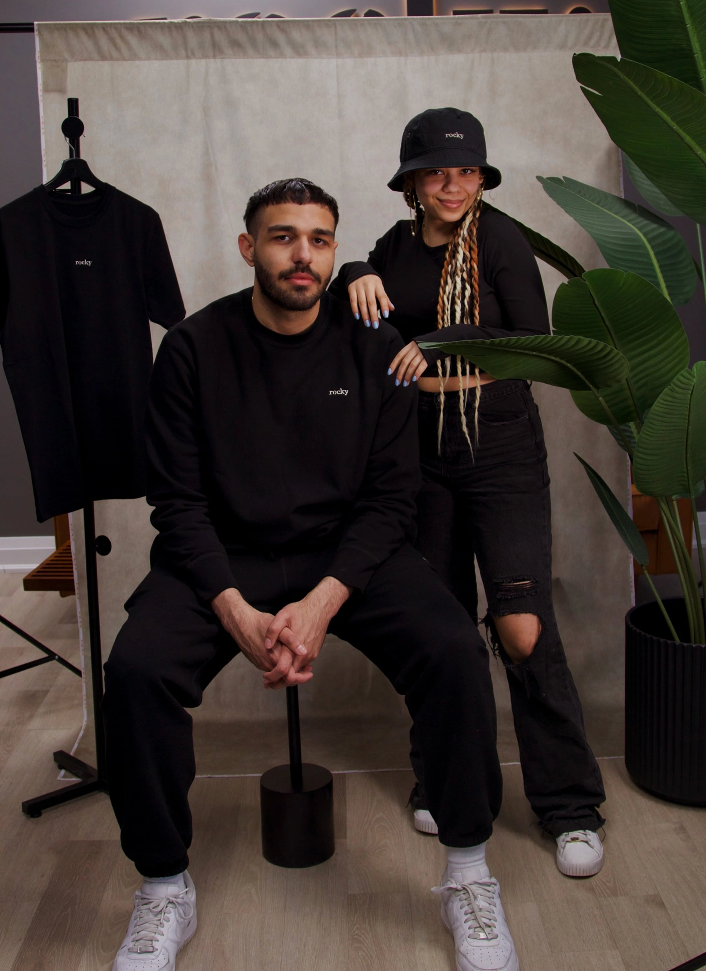 Person with curly hair wearing black top with green plants background